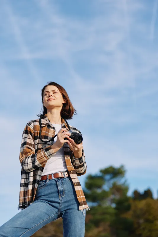 Woman photographer outdoors with camera, wearing a plaid shirt and jeans, standing under a clear blue sky among trees, capturing a candid outdoor moment.