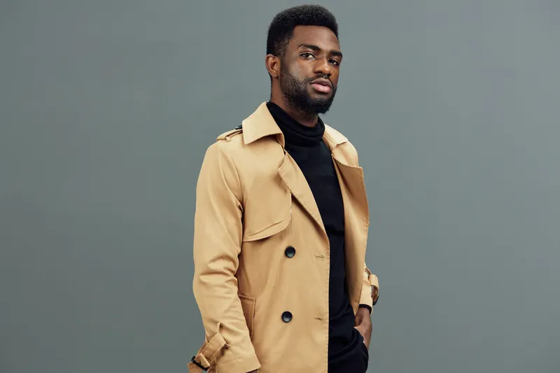 Stylish African man in a beige trench coat and black turtleneck, exuding confidence against a gray background Fashionable, contemporary outfit choice for autumn or winter