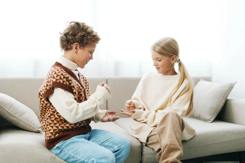 Two children sit on a soft sofa, sharing a quiet moment as one demonstrates a small craft while the other watches with curiosity in a warm, cozy living space.