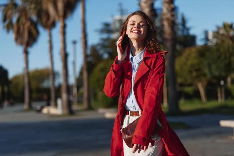 Young woman with curly red hair enjoys a sunny day outdoors while talking on the phone and wearing a red trench coat and white pants.