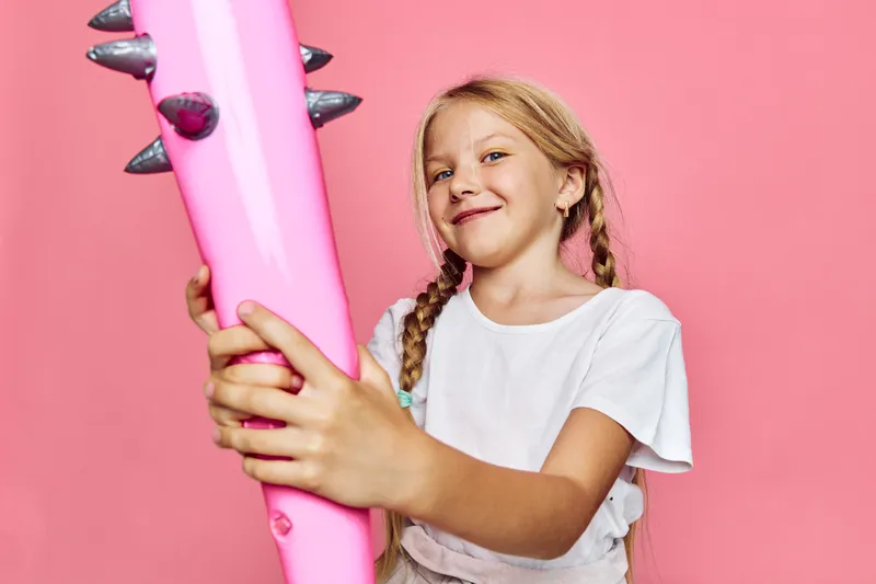 Happy young girl with braided hair holds an inflatable pink bat with spikes standing against a plain pink background. Studio shot with playful and fun concept.