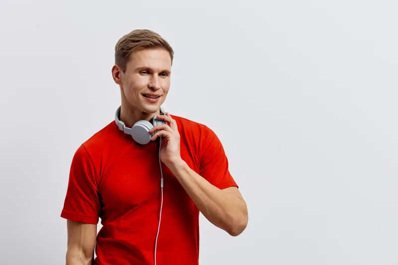 Smiling man with headphones, casual red t-shirt, listening to music or a podcast, standing against a plain white background, studio shot. Lifestyle, leisure, relaxation concept.