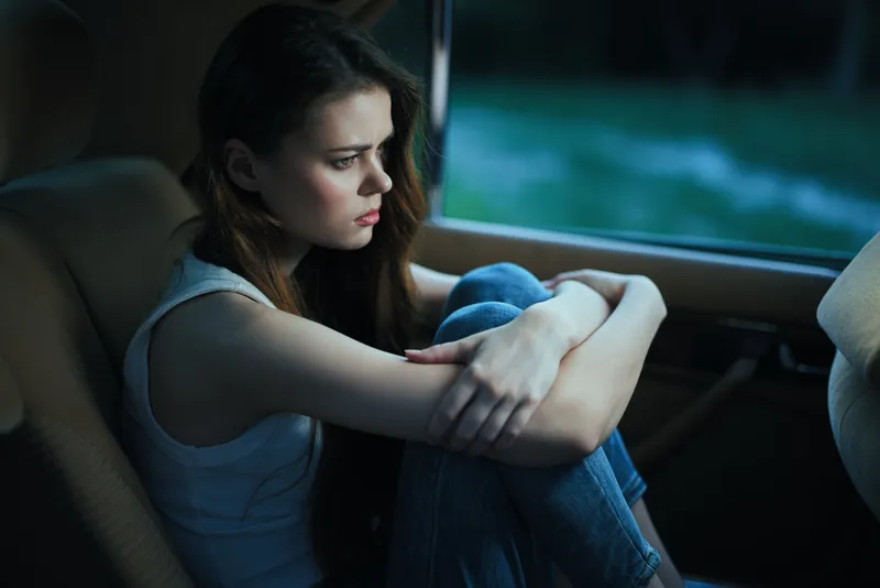 Thoughtful young woman in a car, wearing a white tank top and jeans, expressing sadness against a moody, dark green background.