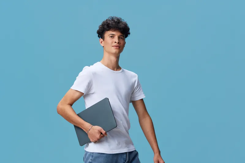 young man with laptop wearing casual white t-shirt stands confidently against blue background showing relaxed posture and modern style, youthful technology concept