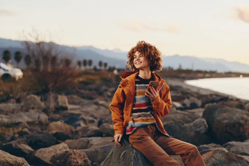 Woman lifestyle outdoors by rocky coast, sunset light, golden evening, smiling curly woman in casual jacket and striped shirt, relaxed pose on stones, gentle breeze, sunlit natural soft warm colors