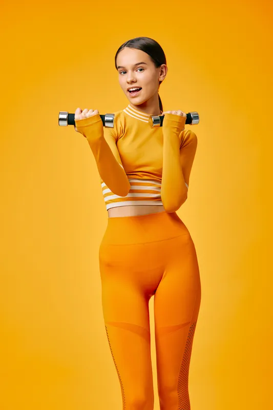 Young woman in bright orange activewear holds a dumbbell in a studio portrait, smiling confidently while demonstrating fitness strength and athletic posture against a seamless yellow background.