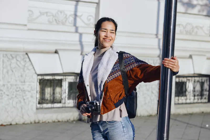 Woman with camera on a sunny street, smiling and leaning on a pole in a casual jacket and scarf, urban backdrop, daytime photography, candid lifestyle shot for street style and travel