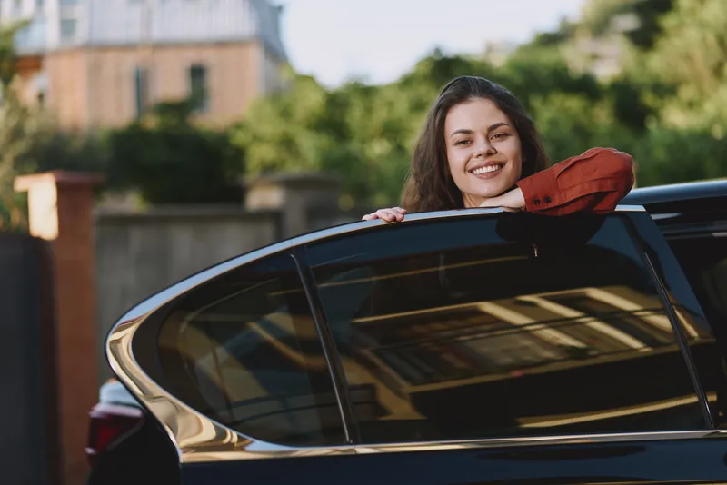Young woman leans from a sleek car window, smiling brightly on a sunlit outdoor street, casual red sleeve fluttering, relaxed confident mood with lush greenery in the background