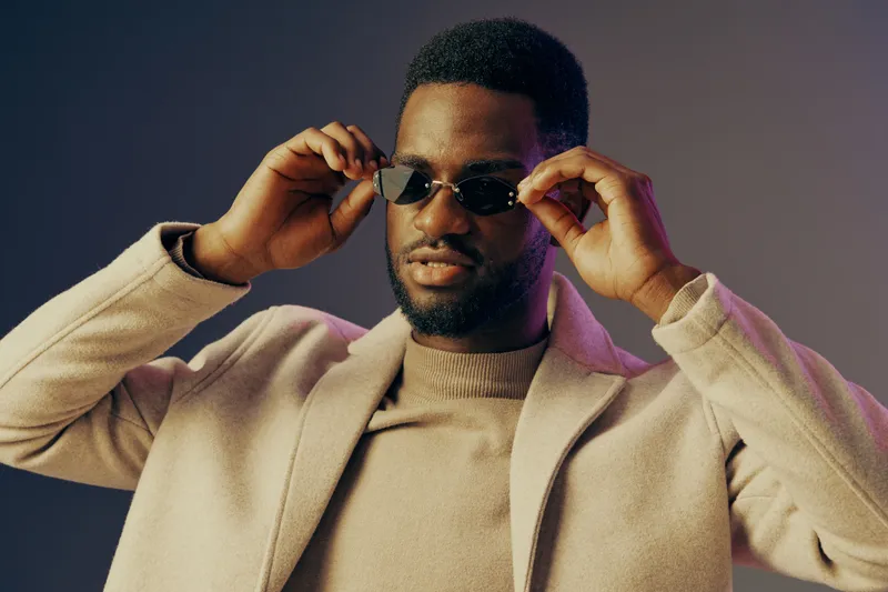 Stylish African male model wearing sunglasses and a beige coat, exuding confidence against a soft gradient background, perfect for fashion and lifestyle themes