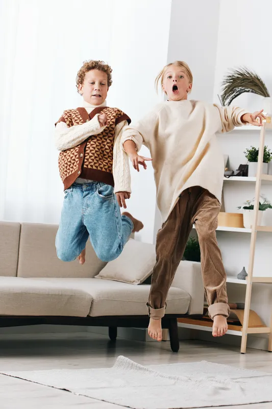 Two children jump mid air in a bright living room, capturing energetic movement, playful youth, and a candid moment of carefree joy indoors. The scene emphasizes dynamic pose and cozy light.