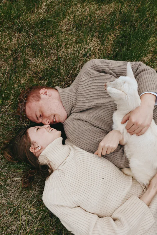 Couple lying on grass, smiling, holding a white goat kid between them. Cozy sweaters and soft light create a warm, intimate lifestyle moment outdoors, highlighting connection, care, and nature.