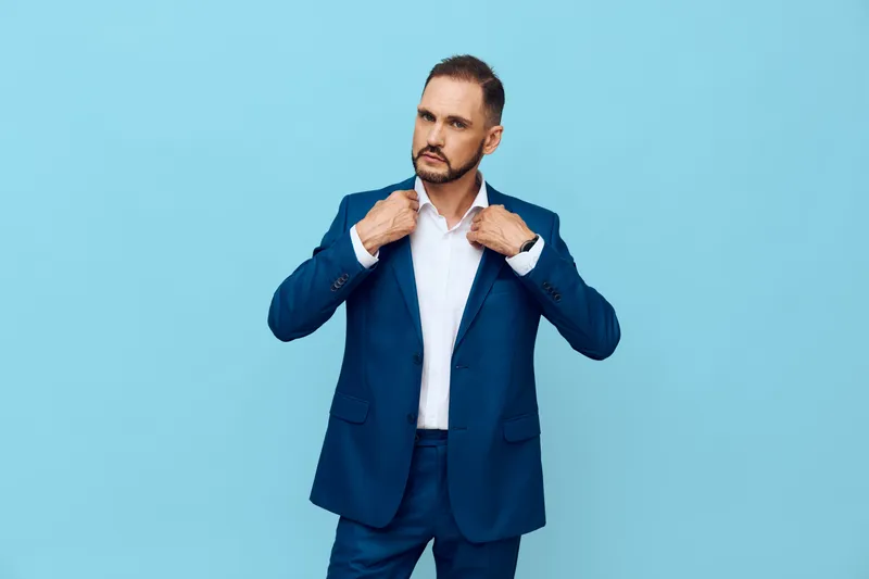 A focused male business professional adjusts his suit against a solid blue backdrop, conveying confidence, readiness, and calm determination.