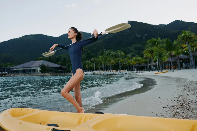 A woman in a navy swimsuit stands on a yellow kayak at the shore, holding two paddles overhead in a joyful pose as gentle waves lap the sand.