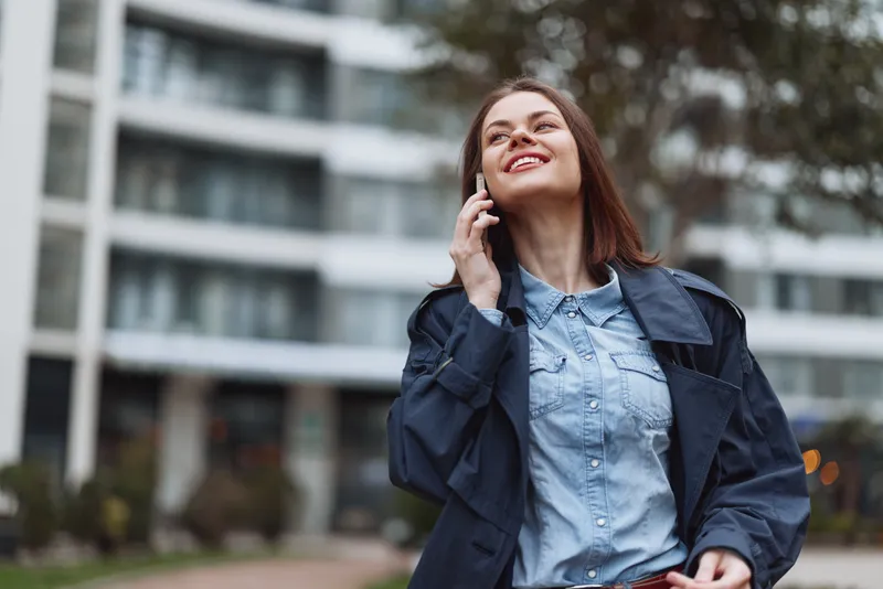 A smiling woman outdoors, talking on a phone with city buildings in the background, wearing a denim jacket and casual shirt.