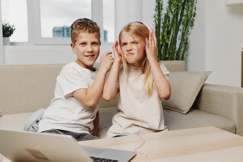 Two children sitting on sofa at home, boy teasing girl with headphones, girl showing annoyed face. Concept of sibling interaction, childhood, family moments and playful behavior indoors.