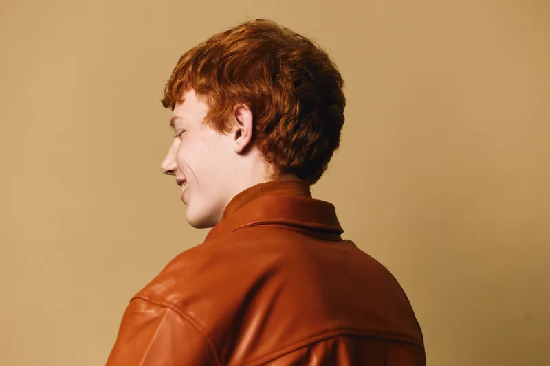 Young man with red hair and fair skin wearing brown jacket stands against beige background looking sideways with relaxed expression for casual portrait.