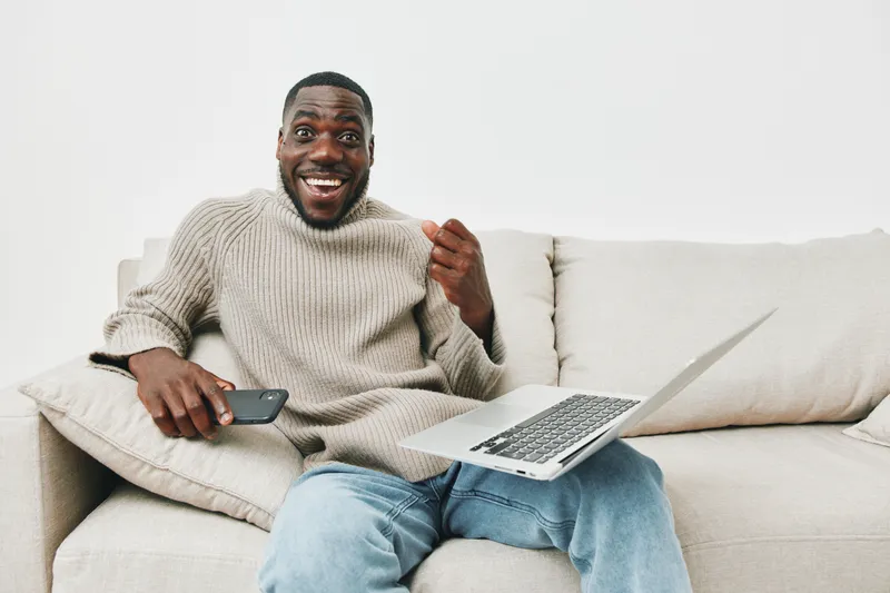 Happy young Black man using smartphone and laptop at home, engaged in remote work with a joyful expression, casual clothing, comfortable setting, natural light