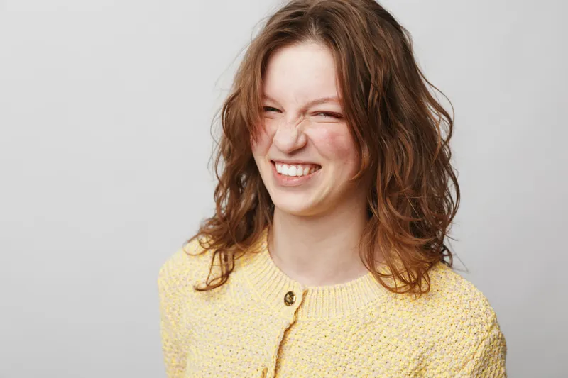 Young woman with curly brown hair wearing yellow knitted sweater smiles with eyes closed on light grey background. Concept of happiness, joy, casual style, and natural beauty portrait.
