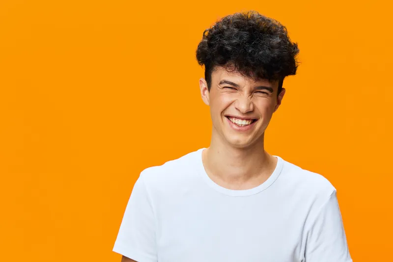smiling young man with curly hair wearing white shirt against vibrant orange background, expressing happiness and carefree positive energy in studio setting
