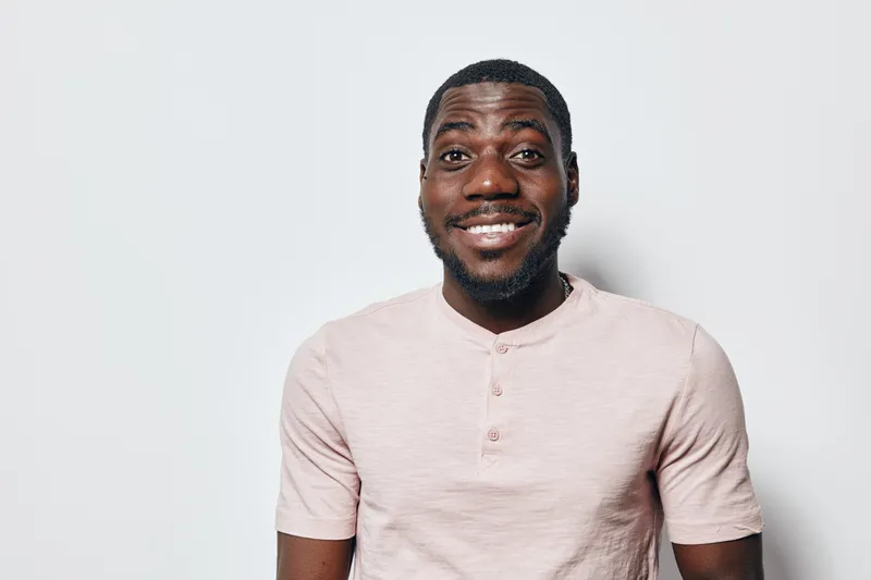 African American man with dark skin smiling and wearing light pink shirt standing against white background. Portrait of happy black male with beard and short hair looking at camera.