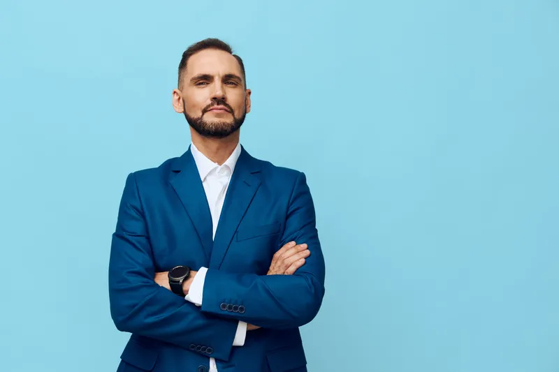 A confident business professional stands with arms crossed against a solid blue backdrop, projecting focus, strength, and readiness for strategic leadership.