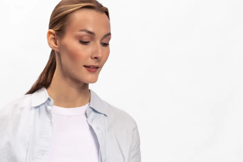 Portrait of a calm young woman with light skin and brown hair looking down. She wears a white shirt and a light blue overshirt against a plain white background. Concept for serenity and contemplation.