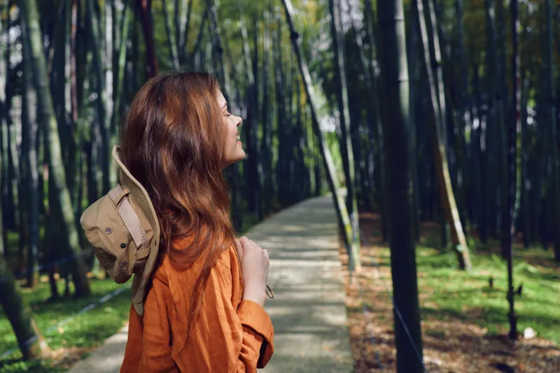 Woman in orange jacket hiking on a forest path through tall bamboo, portrait side view with hat on her back, enjoying nature walk and serene outdoor atmosphere