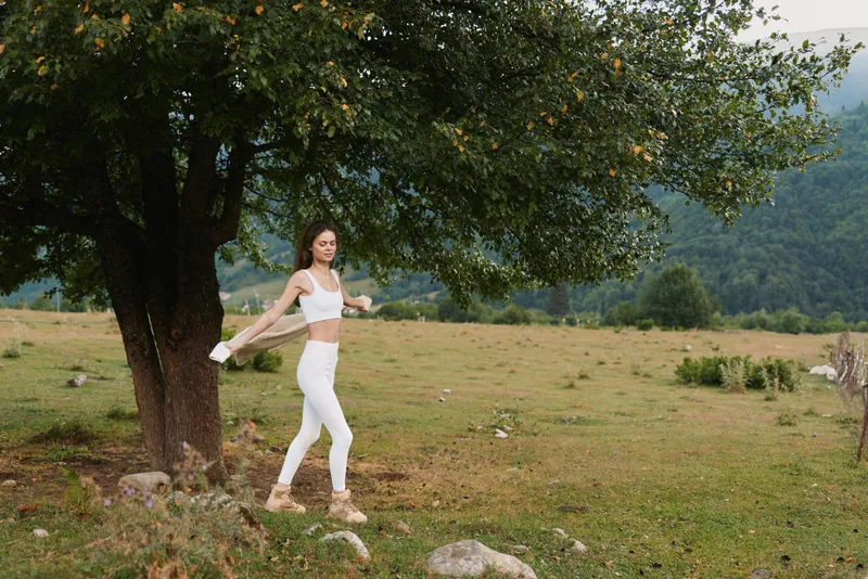 Young woman in white sportswear walking near a large tree in a green field with mountains in the background. Concept of lifestyle, fitness, and nature relaxation.