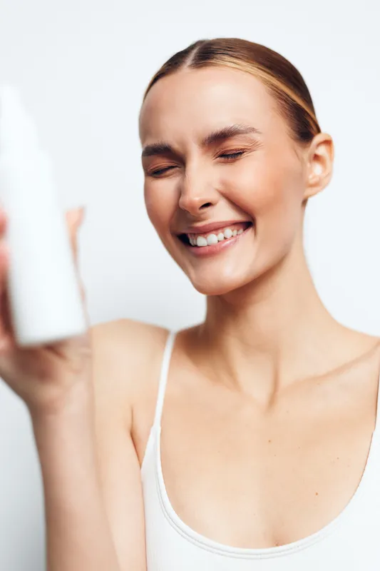 Smiling woman with clear skin holding white cosmetic bottle close to face on white background. Concept of skincare, beauty, wellness, and natural care in bright soft lighting.