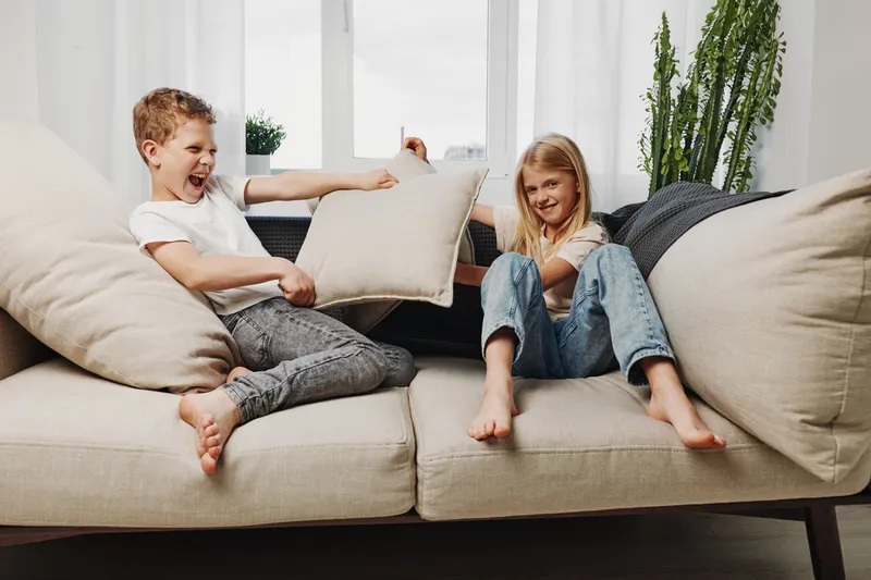 Two playful children having a pillow fight on a beige sofa in a bright living room with casual clothes and natural light.