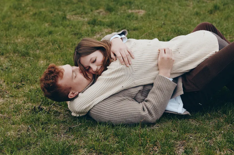 Couple embrace on grass outdoors, sharing warmth and love in a tender moment. Cuddling in cozy sweaters under soft daylight, this lifestyle scene conveys romance, calm, and connection. Natural