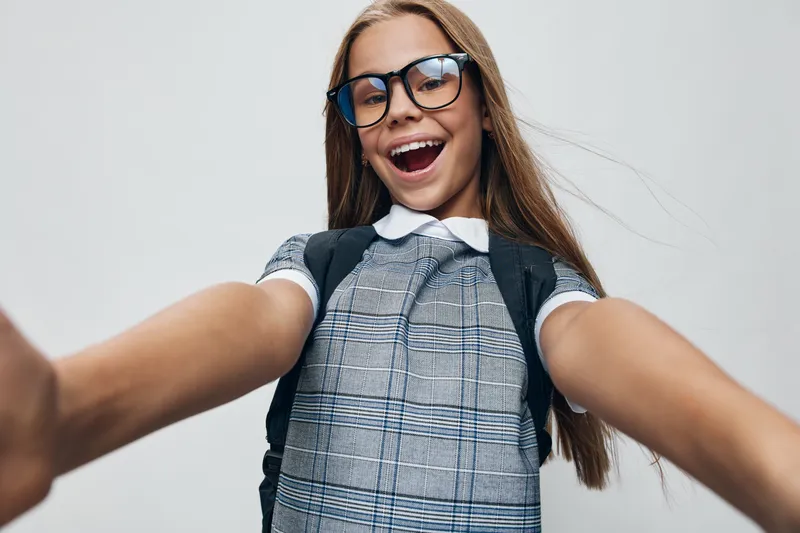 Girl child with glasses smiling and making selfie with outstretched arms against neutral background. Happy schoolgirl in checkered dress and backpack taking photo.