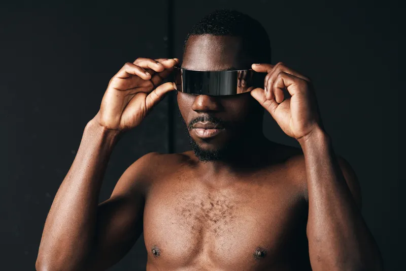 Black man with dark skin and bare chest wearing futuristic visor in creative conceptual visual style. Portrait of confident African ethnicity male with modern technology accessory on black background.