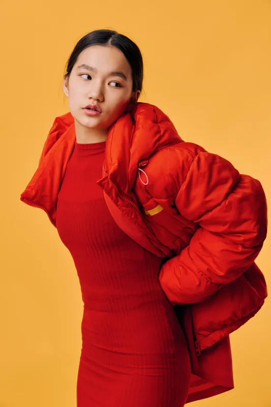 Woman in red dress and red puffer jacket posing on yellow background looking sideways with thoughtful expression, fashion style and casual outfit for autumn or winter season.