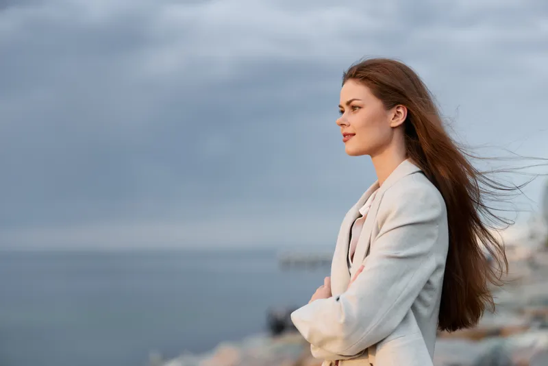 Woman with long brown hair in light jacket thoughtfully looking at sea on windy day, calm expression, natural light, outdoor coastal background, autumn season