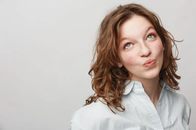 Young woman with curly hair wearing white shirt looks up with thoughtful expression on light background. Concept for contemplation, decision making, and casual lifestyle.