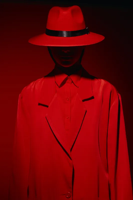 Woman wearing an oversized red blazer and wide-brimmed hat in dramatic lighting standing against a deep red background. Concept of fashion creativity and bold style.