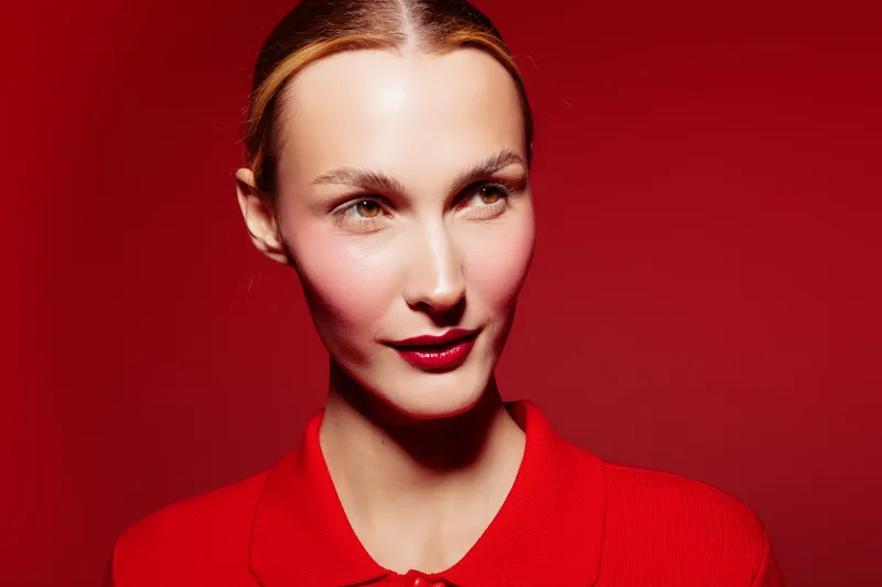 Portrait of a confident woman with red lipstick and red shirt against a red background. The image highlights beauty, fashion, and bold color contrast in studio lighting.