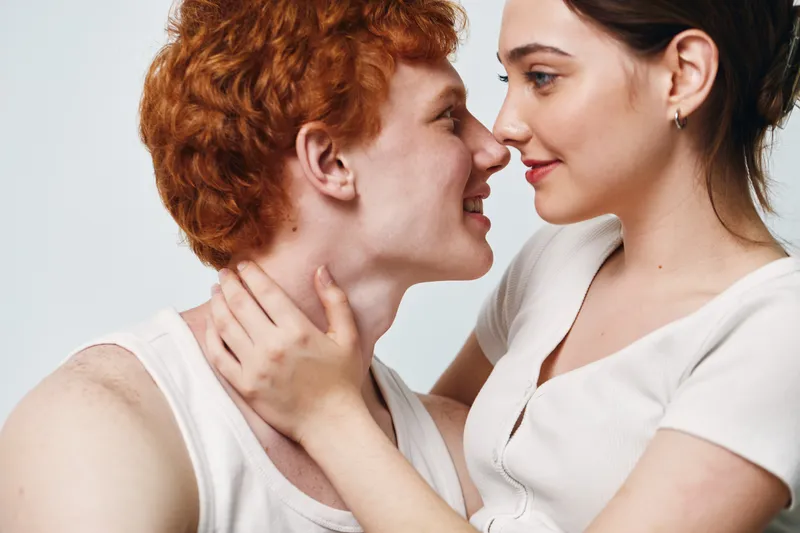 Couple, young man and woman looking at each other, smiling and touching in close intimate moment with soft natural light on white background, affection and connection.