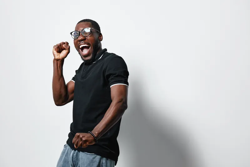 African American man with dark skin wearing glasses and black polo shirt celebrating success with raised fist and joyful expression on white background in studio.