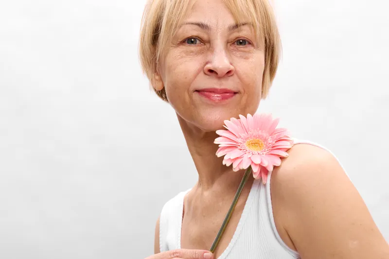 Woman senior smiling portrait flower gerbera pink closeup headshot skincare concept, beauty wellness and selfcare, calm youth, soft studio light, white background