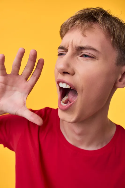 Young man shouting into his cupped hand on a bright yellow background, wearing a red shirt, capturing a dynamic, energetic vocal moment with expressive intensity.