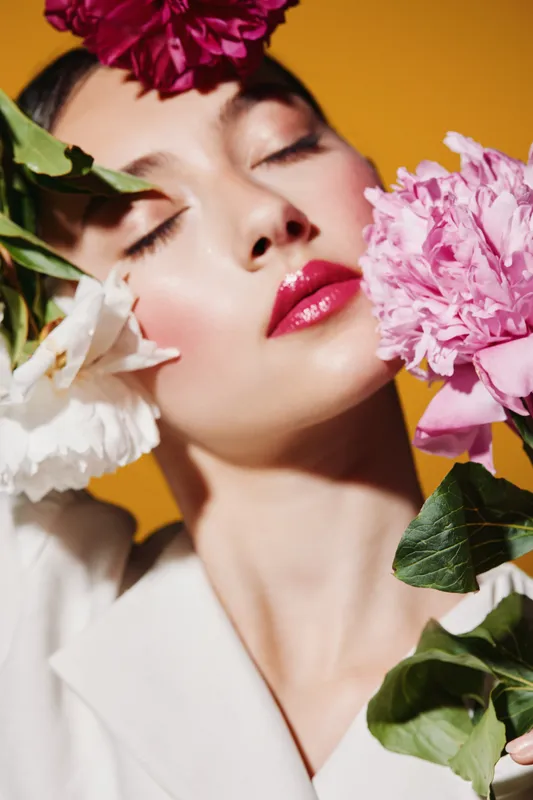 Portrait of a creative beauty model with glossy red lips and blooming flowers around closed eyes. Fashion makeup, expressive floral concept on orange background.