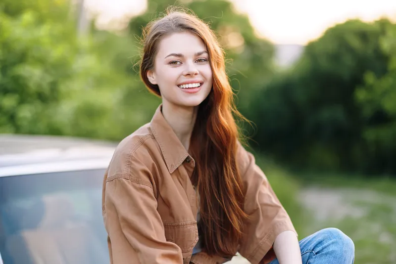 Young woman smiling joyfully in a casual outfit, enjoying nature outdoors in a lush green background during a sunny day, embodying happiness and freedom.