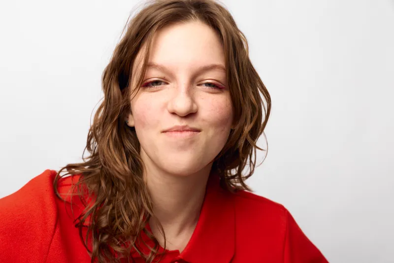 Young woman with wavy brown hair wearing a red shirt smiles subtly against a white background. Casual portrait with natural expression and soft lighting for lifestyle concept.