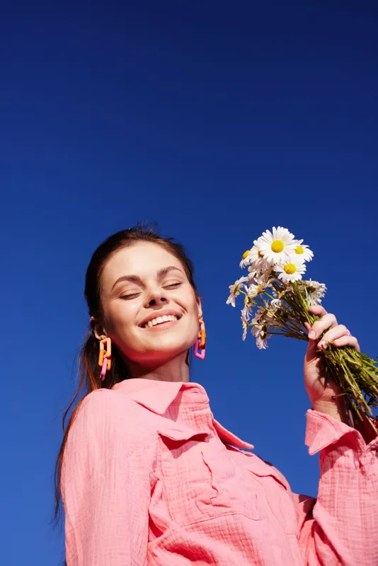 Young woman in pink shirt holding daisies, smiling against a bright blue sky, representing happiness and springtime vibes