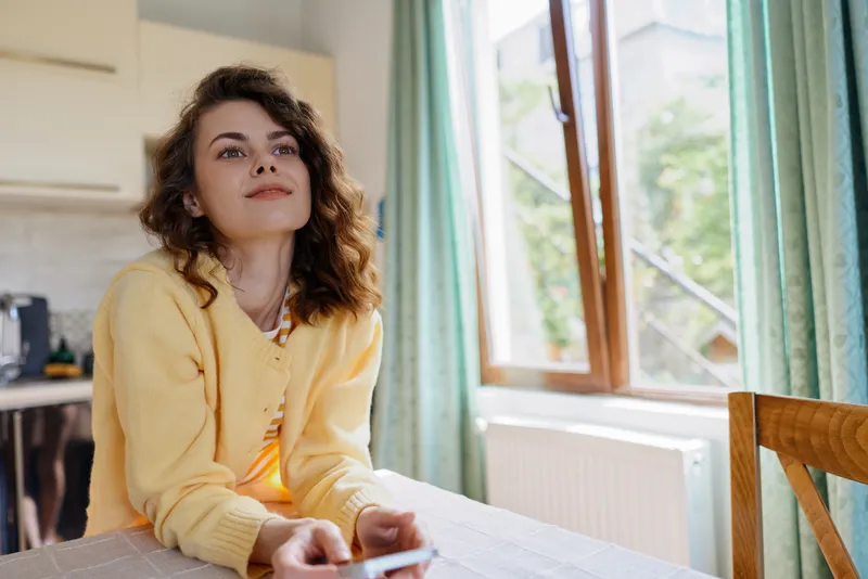 Young woman sits at a home table in a yellow cardigan, gazing upward with a soft smile as sun streams through a window, conveying a relaxed morning mood.