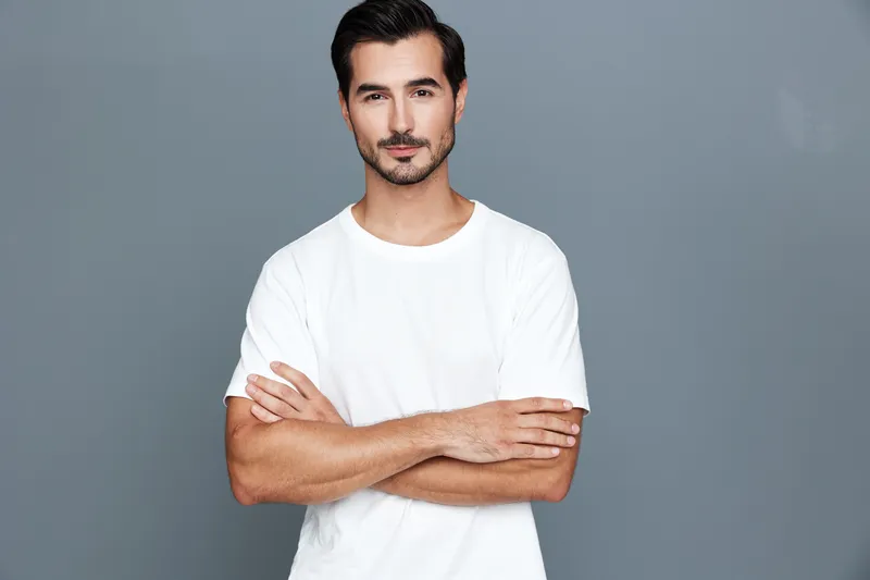 Confident young caucasian man in casual white t-shirt standing with arms crossed, looking at camera against a solid gray background. Fashion portrait concept.