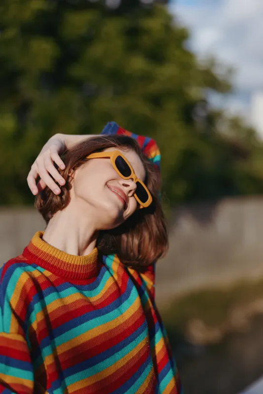 woman smiling in a colorful striped sweater and yellow sunglasses, short hair leaning back in sunlight outdoors, casual outfit, playful lifestyle portrait in a leafy park setting