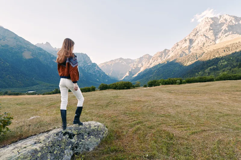 Woman standing on rock wearing sweater and boots, back view looking at mountain landscape and meadow during hiking trip, nature travel and scenic solitude in alpine outdoors.